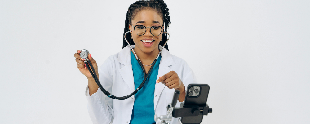 Smiling physician in white coat and teal scrubs holding up a stethoscope while recording a health tip on a phone tripod
